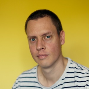 A portrait of James against a yellow background, wearing short hair and a back and white striped t-shirt.